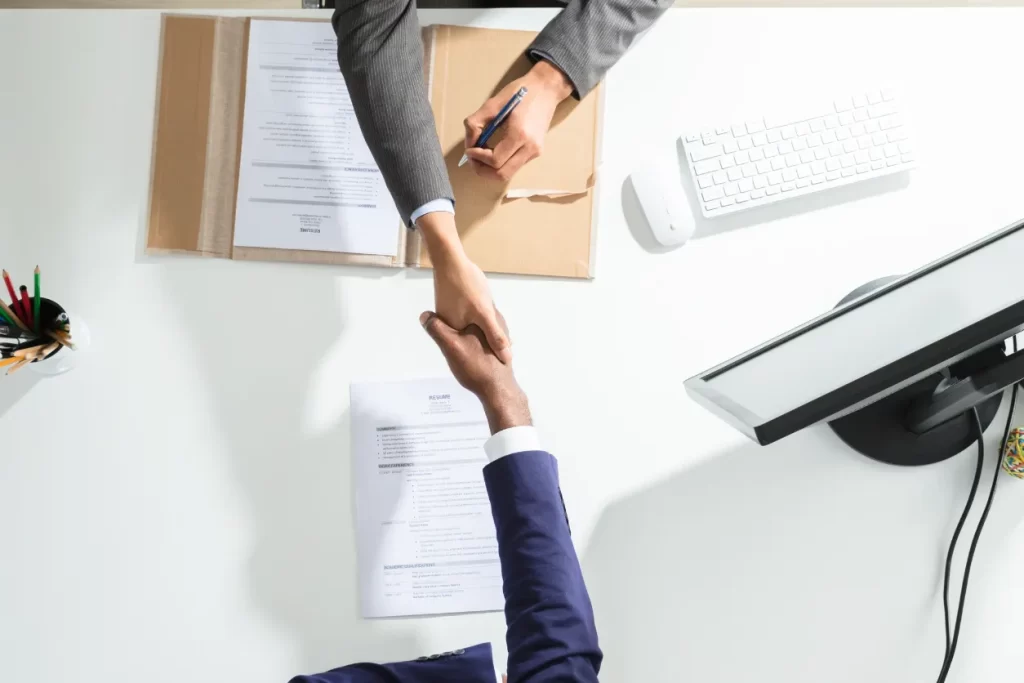 Top-down view of a business handshake across a desk with resumes at Moving Heads Recruitment Agency